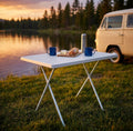 Tavolino pieghevole in acciaio e PVC bianco allestito in riva al lago al tramonto con tazze smaltate e pane.