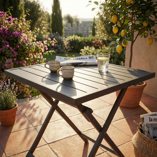 Dettaglio del tavolo pieghevole Alabama illuminato dalla luce solare pomeridiana, con giornale e tazze in ceramica artigianale.