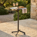 Tavolo alto Vega in acciaio taupe allestito per un aperitivo all'aperto con calici e stuzzichini.