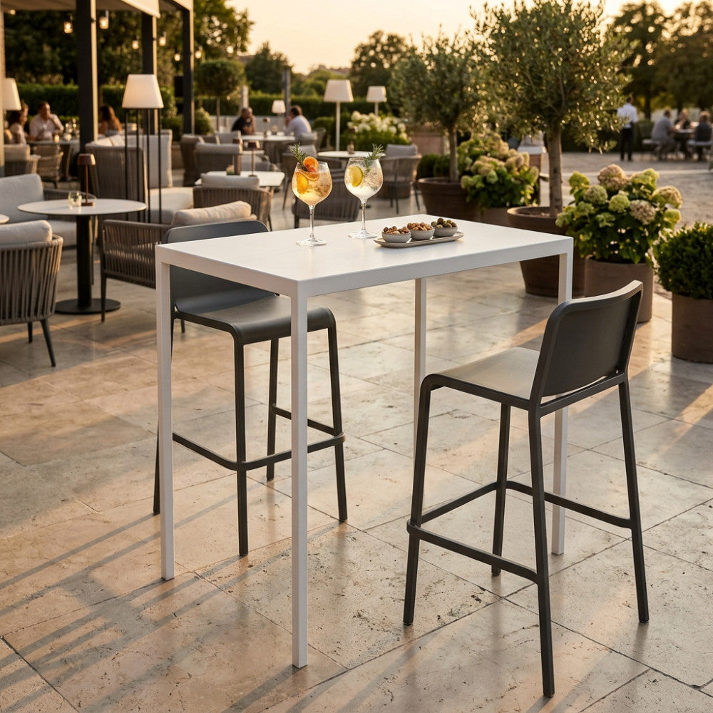 Tavolo alto da giardino moderno in metallo bianco allestito in un patio esterno soleggiato con due sgabelli scuri e bevande rinfrescanti.