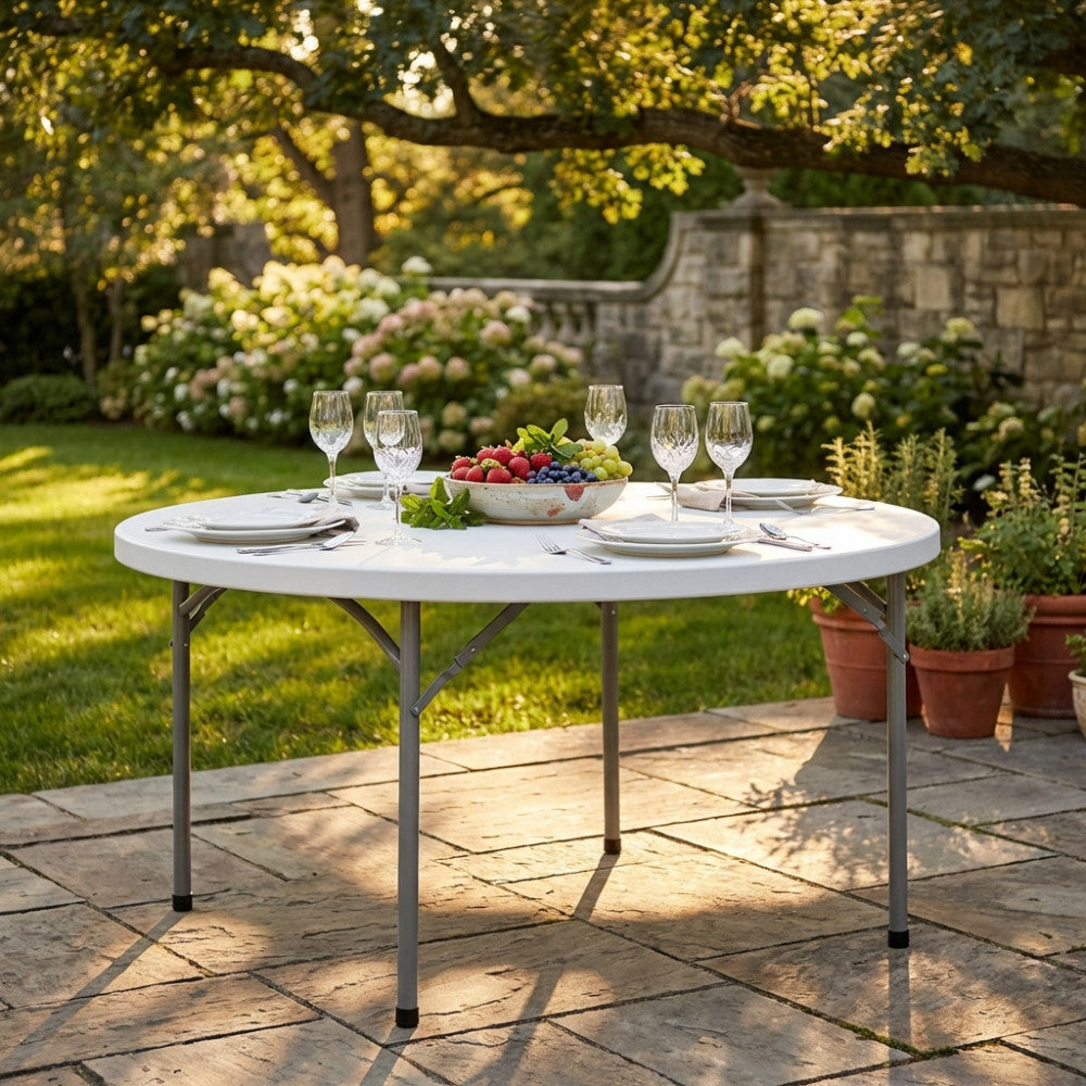 Tavolo da catering rotondo in metallo bianco allestito in un giardino lussuoso con frutta e calici di cristallo.