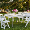 Tavolo da catering rotondo allestito per un ricevimento all'aperto con sedie bianche pieghevoli.
