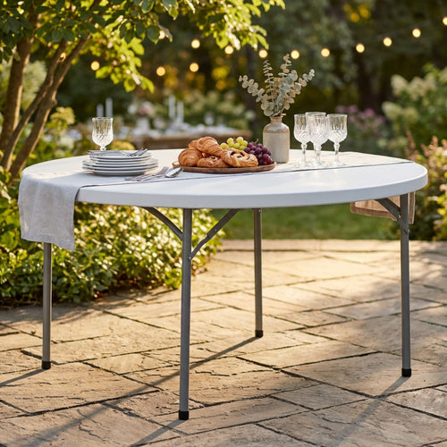 Tavolo rotondo da catering allestito in un giardino lussuoso con tovaglia in lino, stoviglie eleganti e un vassoio di croissant sotto luci calde.