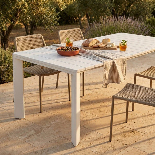 Dettaglio del piano a doghe del tavolo in alluminio bianco decorato con pane, formaggio e bevande fresche alla luce del tramonto.