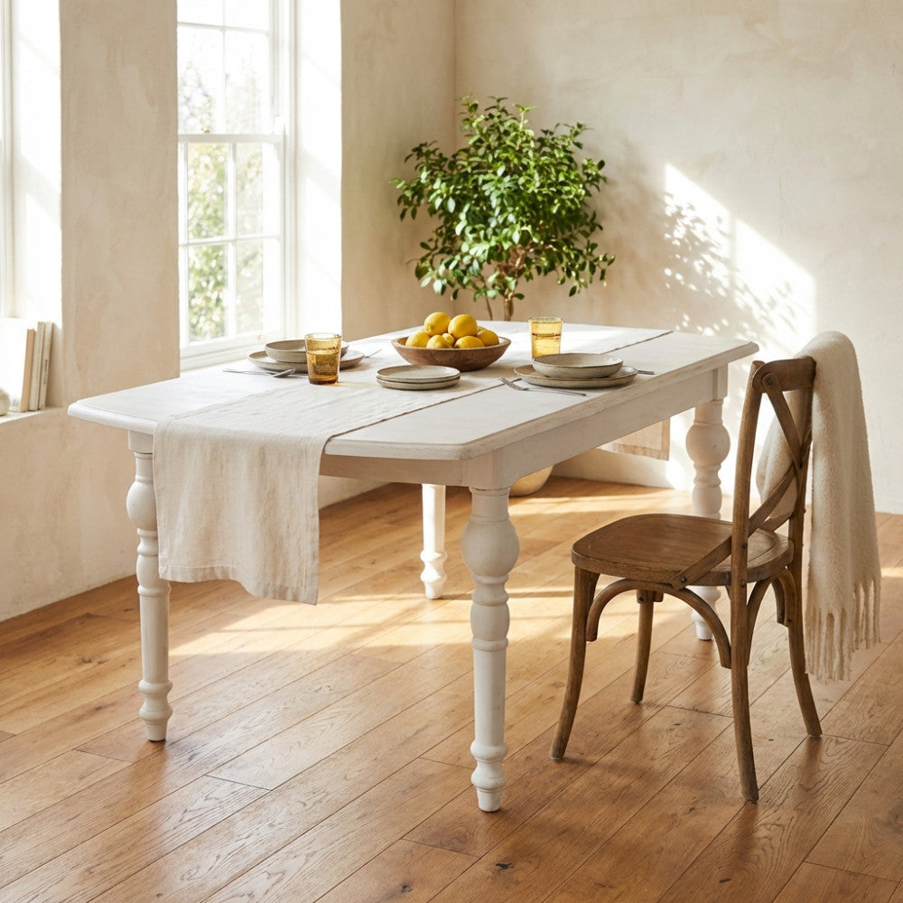 Tavolo da pranzo in legno bianco estensibile, allestito con runner in lino e limoni freschi in un ambiente rustico-chic illuminato dal sole.