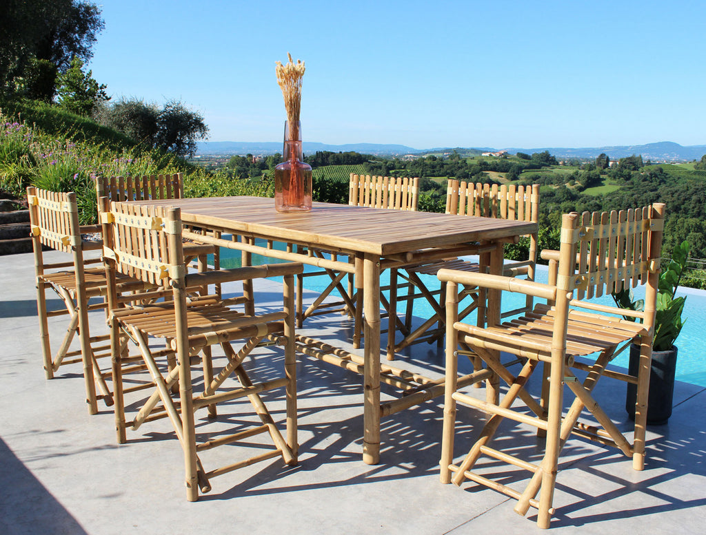 Vista dettagliata del tavolo da giardino in legno Cuba Libre e sedie incorniciati da un paesaggio di campagna panoramico.