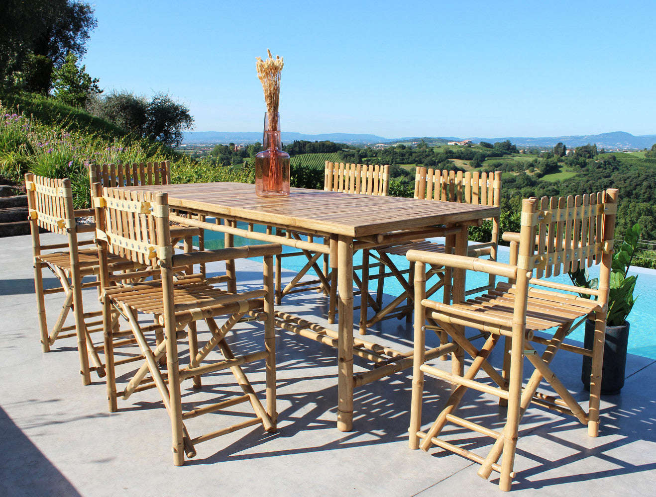 Vista dettagliata del tavolo da giardino in legno Cuba Libre e sedie incorniciati da un paesaggio di campagna panoramico.