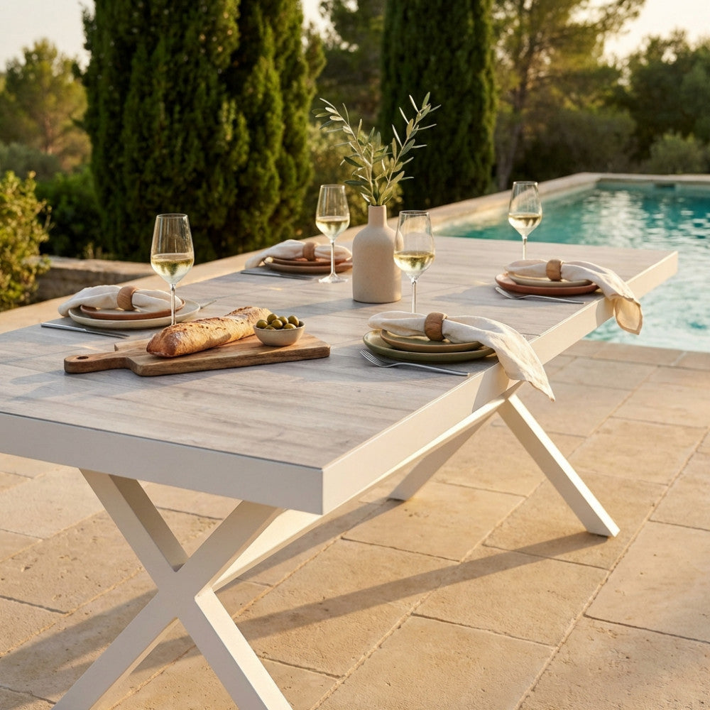 Tavolo outdoor Djerba in gres e alluminio bianco a bordo piscina, decorato con calici di vino e pane rustico.