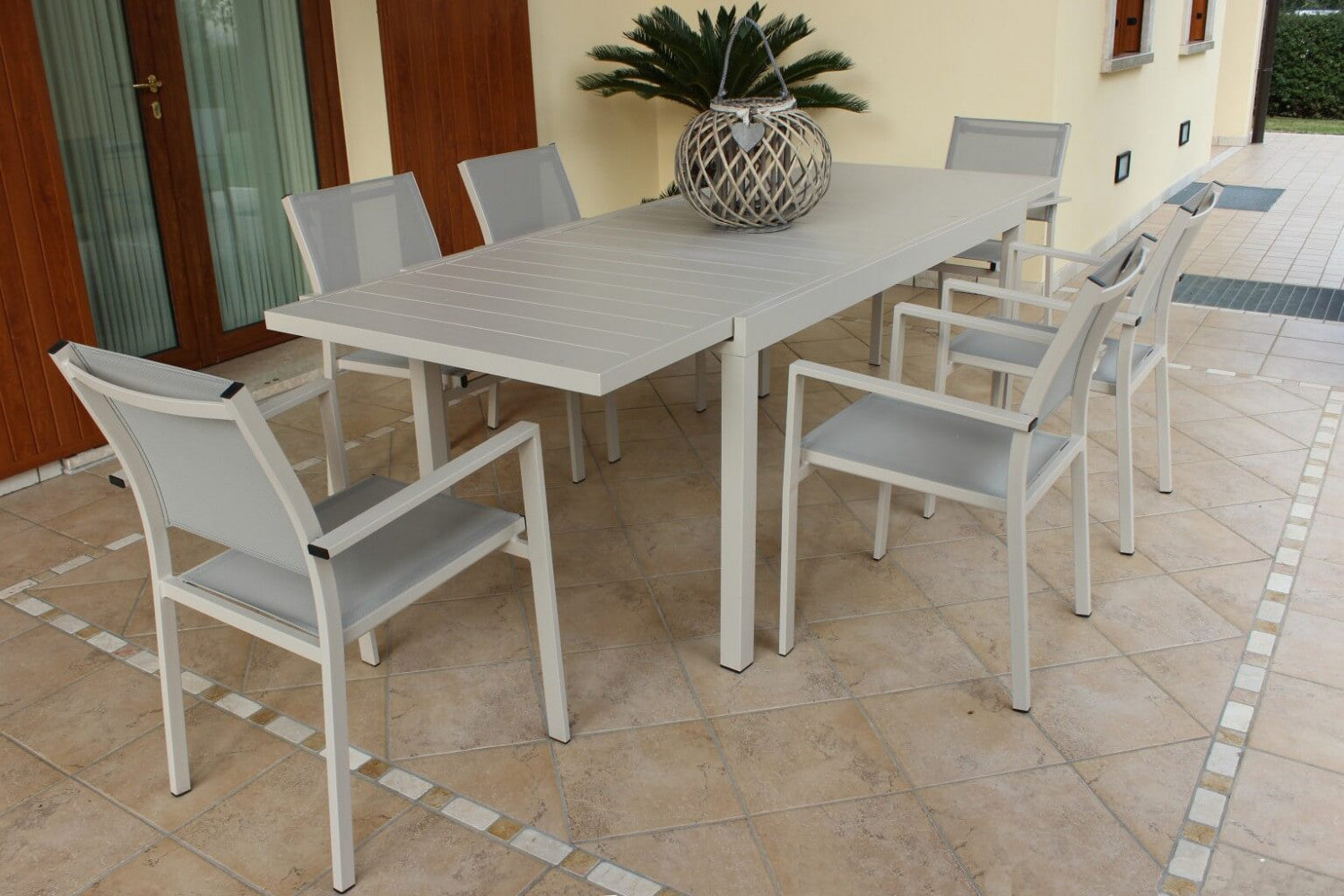 Tavolo Formentera in alluminio tortora ambientato in un patio esterno, circondato da sedie con braccioli.