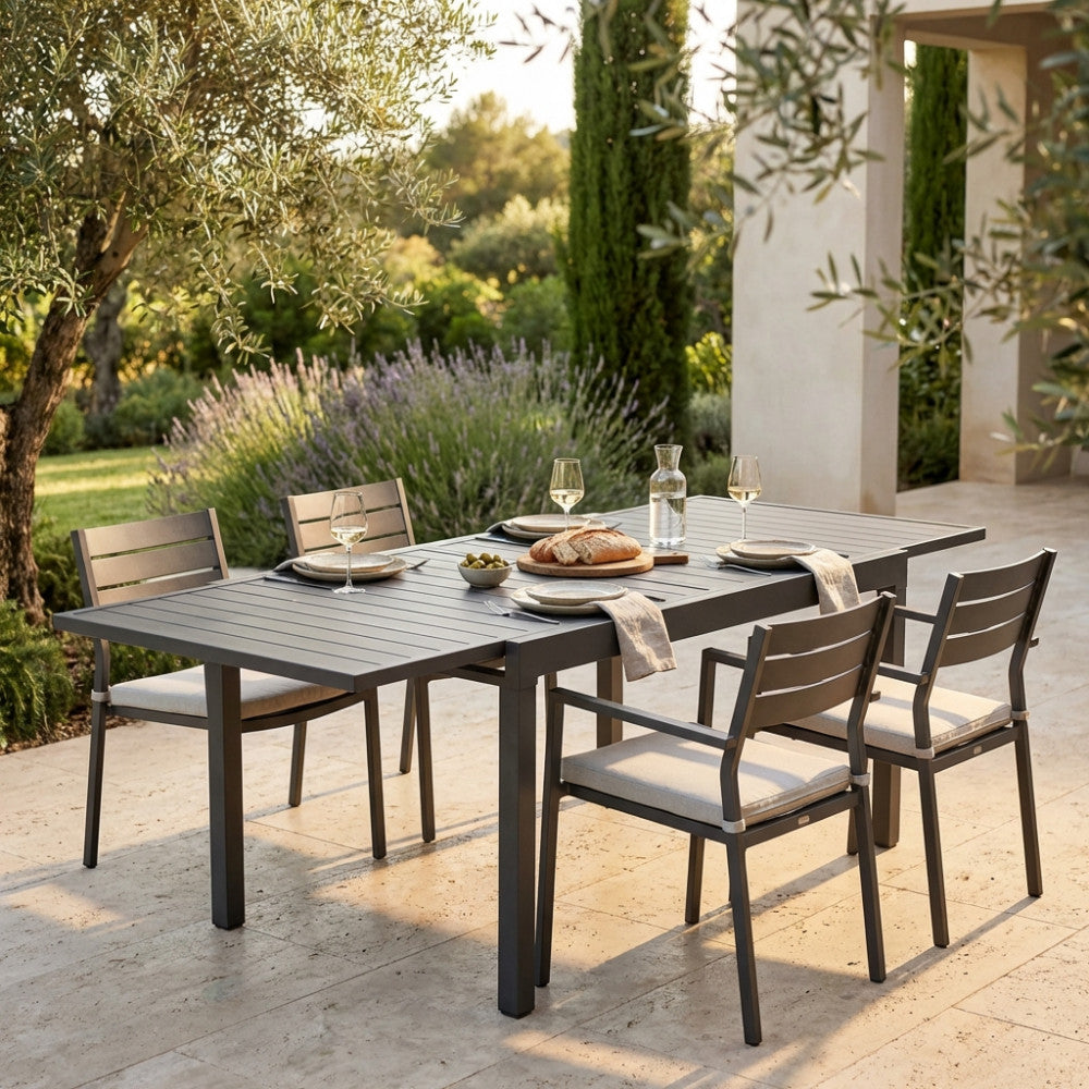 Tavolo da pranzo allungabile Hawaii color taupe, elegantemente allestito per un pasto all'aperto in un giardino lussureggiante, accompagnato da sedie a doghe abbinate.