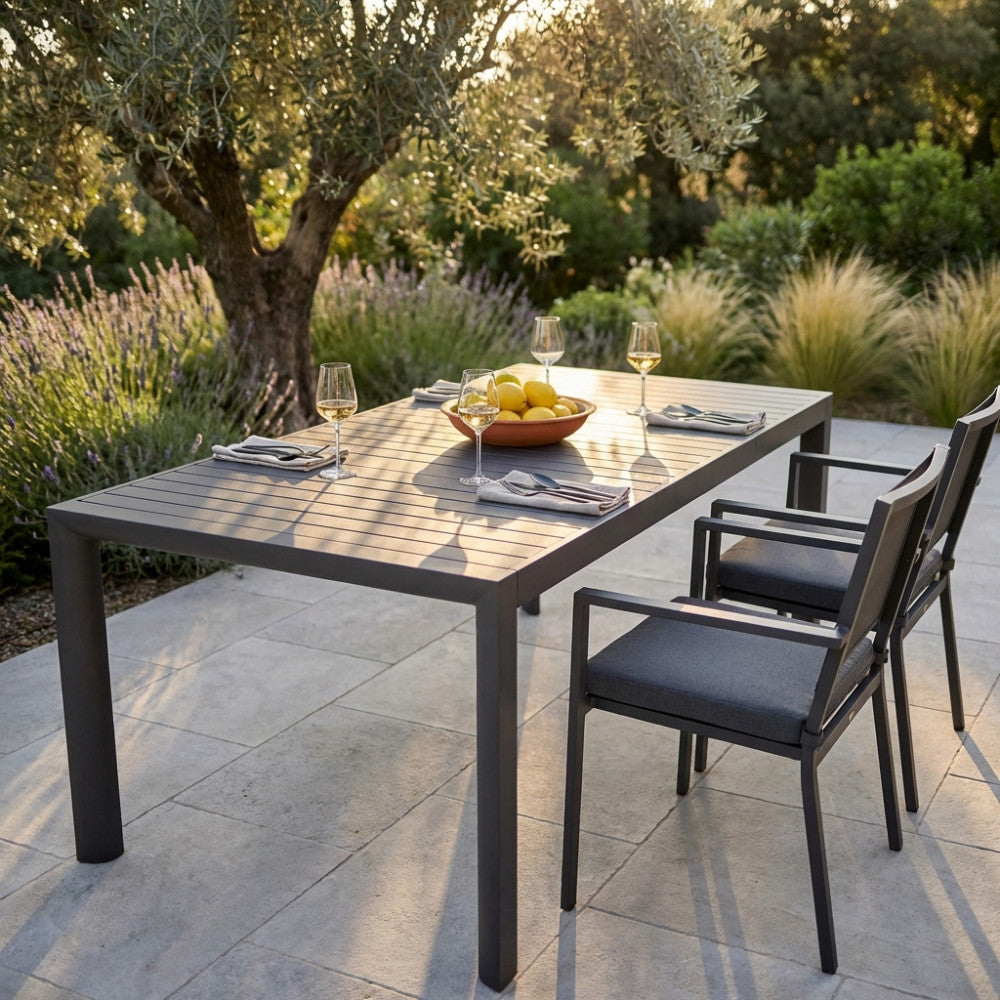 Dettaglio del tavolo Houston in alluminio illuminato dalla calda luce del tramonto, apparecchiato elegantemente per una cena all'aperto in un giardino mediterraneo.