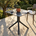 Tavolo da giardino Jarvis 70x70 antracite con piano microforato, allestito su un patio in pietra con pianta e tazzina da caffè.