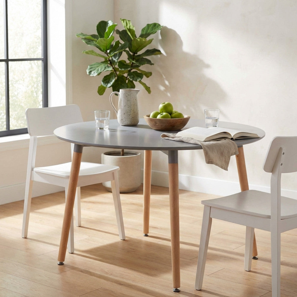 Tavolo da pranzo rotondo grigio Lione con gambe in legno, allestito in una sala luminosa con sedie bianche e un cesto di mele verdi.