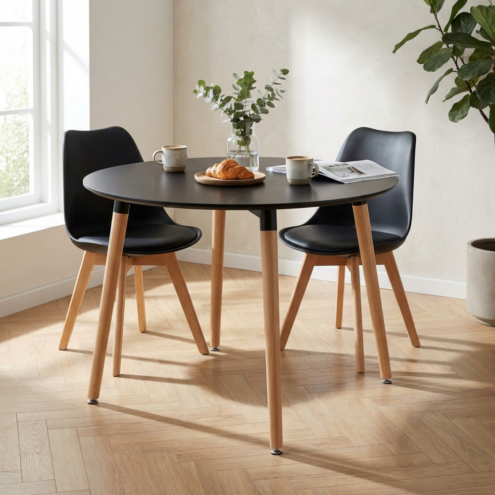 Tavolo da pranzo rotondo nero con gambe in legno, allestito con caffè e croissant, accompagnato da due sedie nere in un ambiente luminoso.