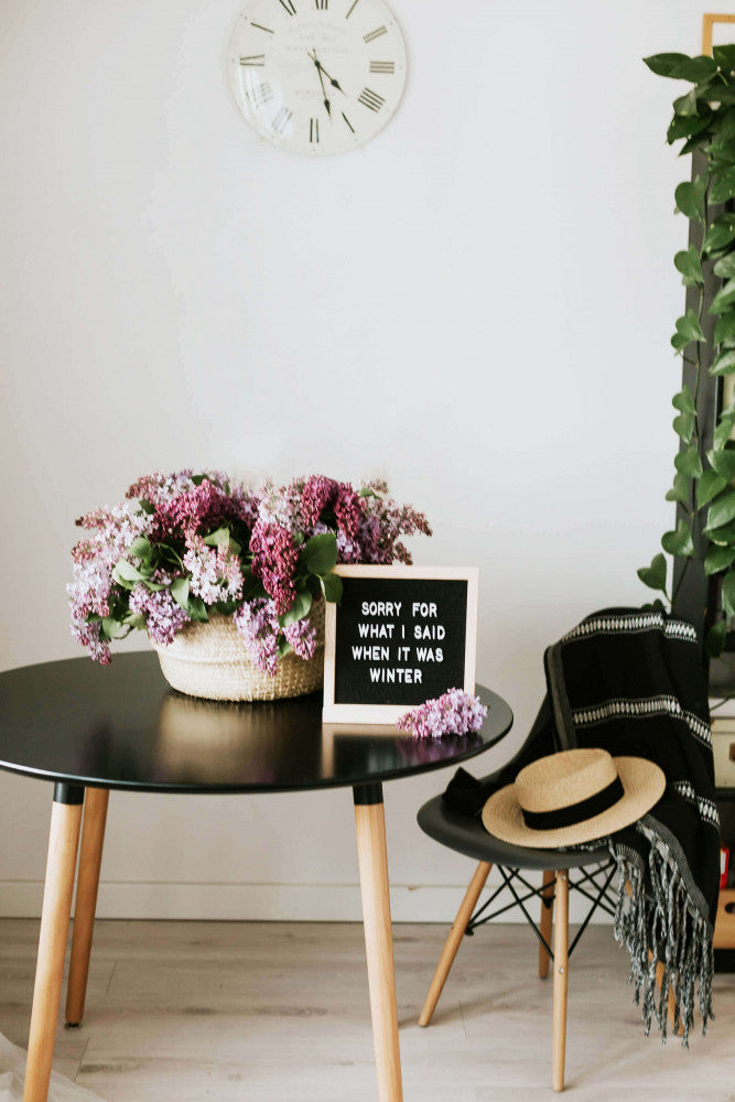 Dettaglio lifestyle del tavolo nero decorato con un cesto di fiori lilla e una lavagna con scritte, accanto a una sedia con cappello.