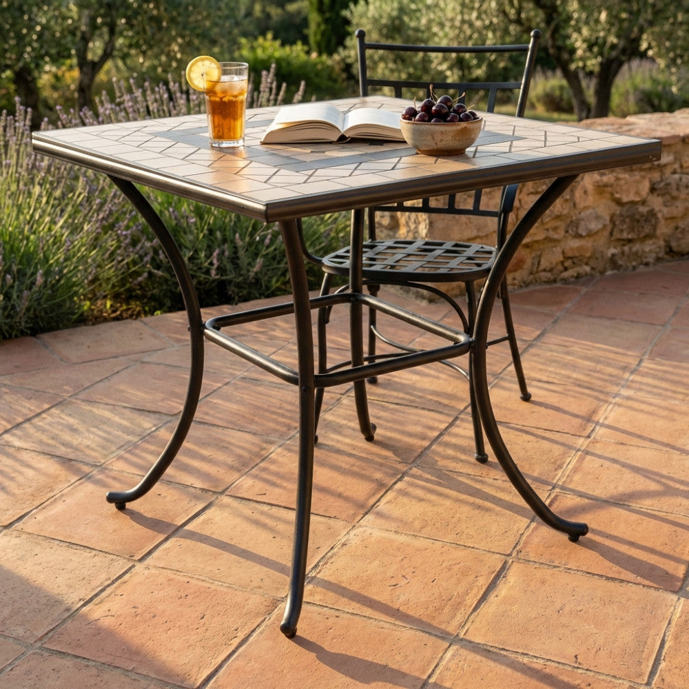 Tavolo da giardino quadrato Nereo in cemento taupe e acciaio, allestito con bevanda e ciliegie su un patio in cotto.