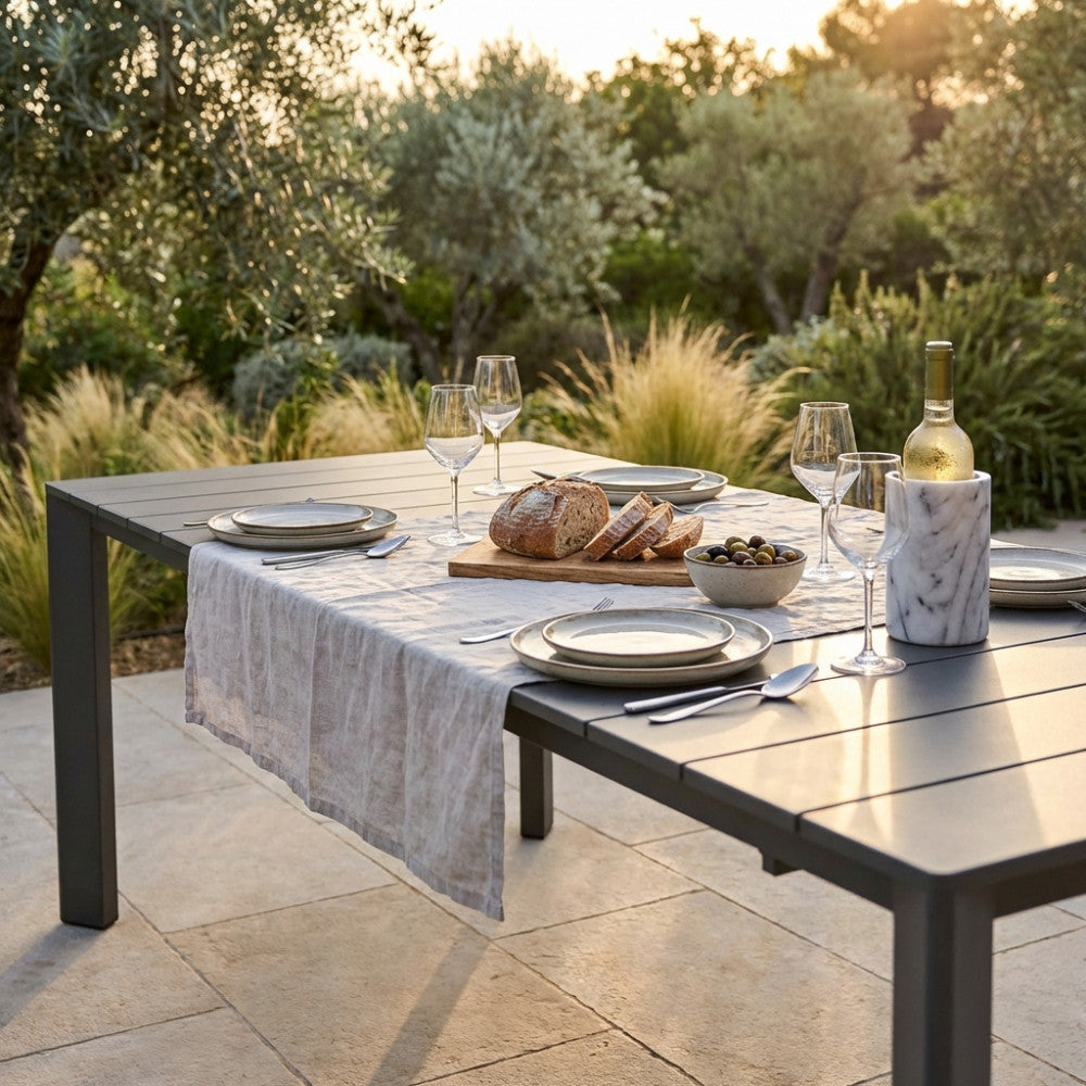 Dettaglio della superficie in alluminio taupe del tavolo Sacramento illuminata dalla luce del tramonto con accessori da tavola eleganti.