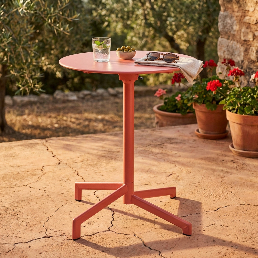 Tavolo da giardino Vega in acciaio rosso corallo ambientato all'aperto con bicchiere e occhiali.