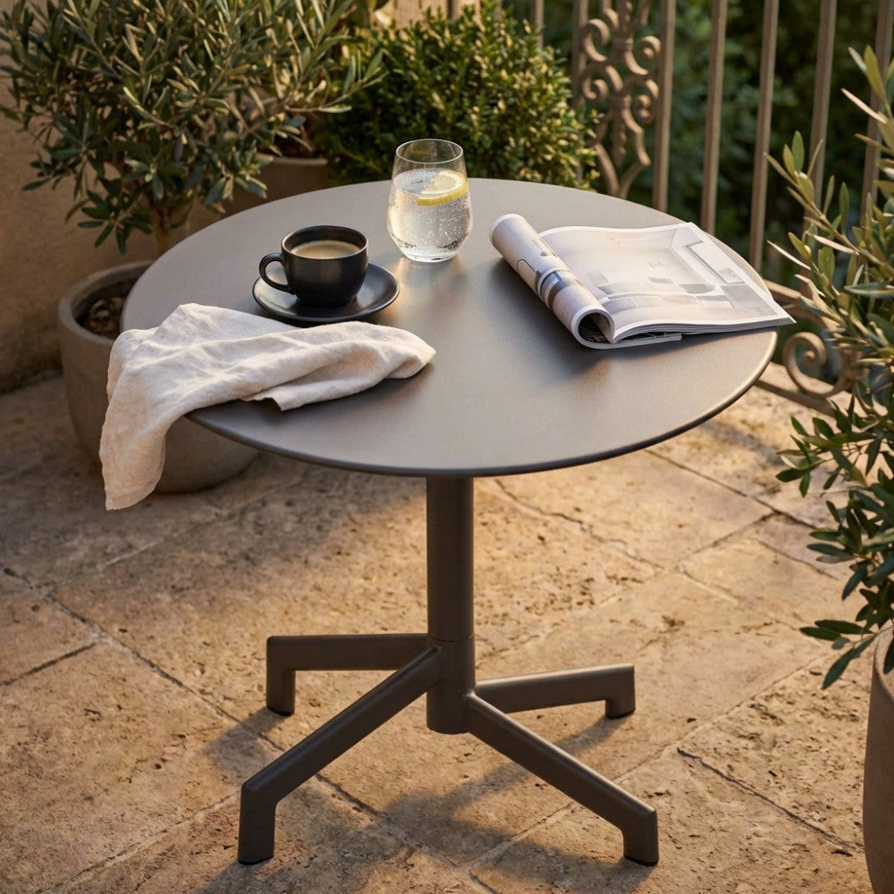 Tavolo da giardino rotondo in acciaio taupe allestito con caffè e rivista su un patio in pietra.