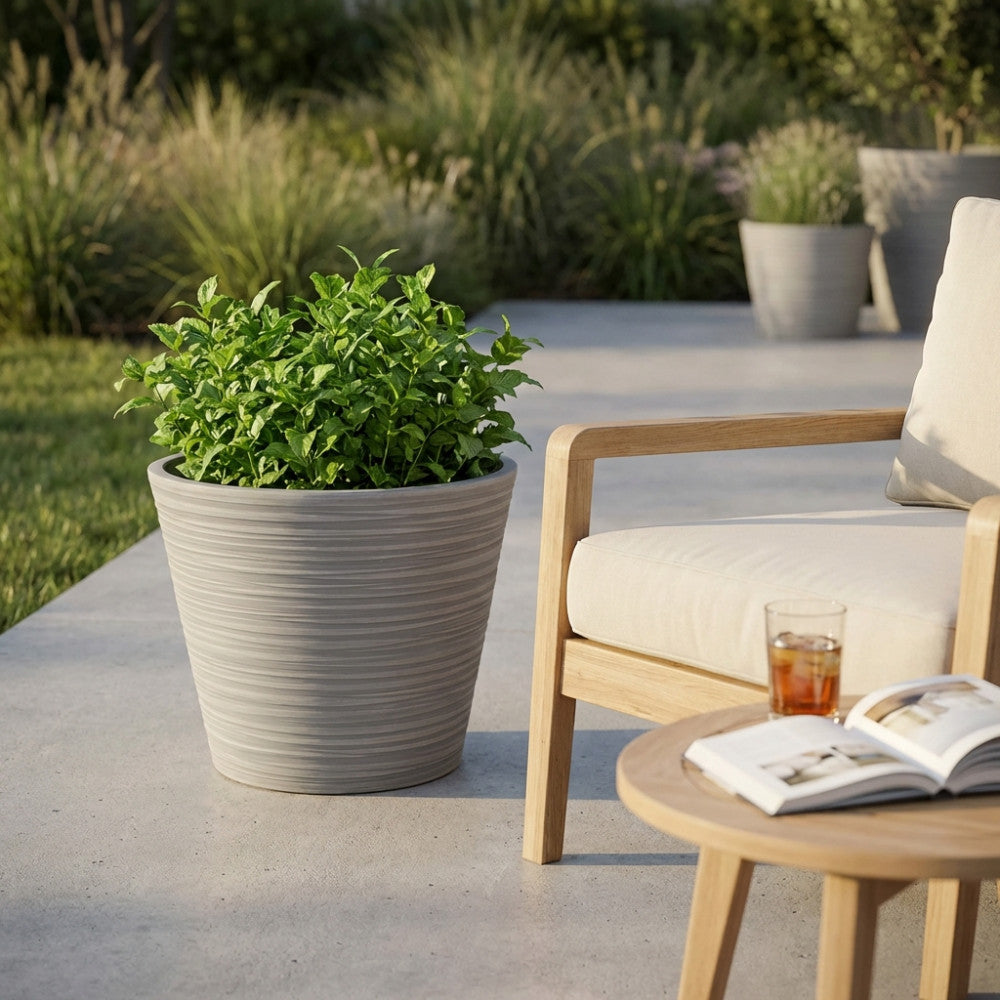Dettaglio del Vaso Camelia color antracite con pianta di menta, situato vicino a una poltrona in legno di teak e un tavolino da caffè in un giardino elegante.