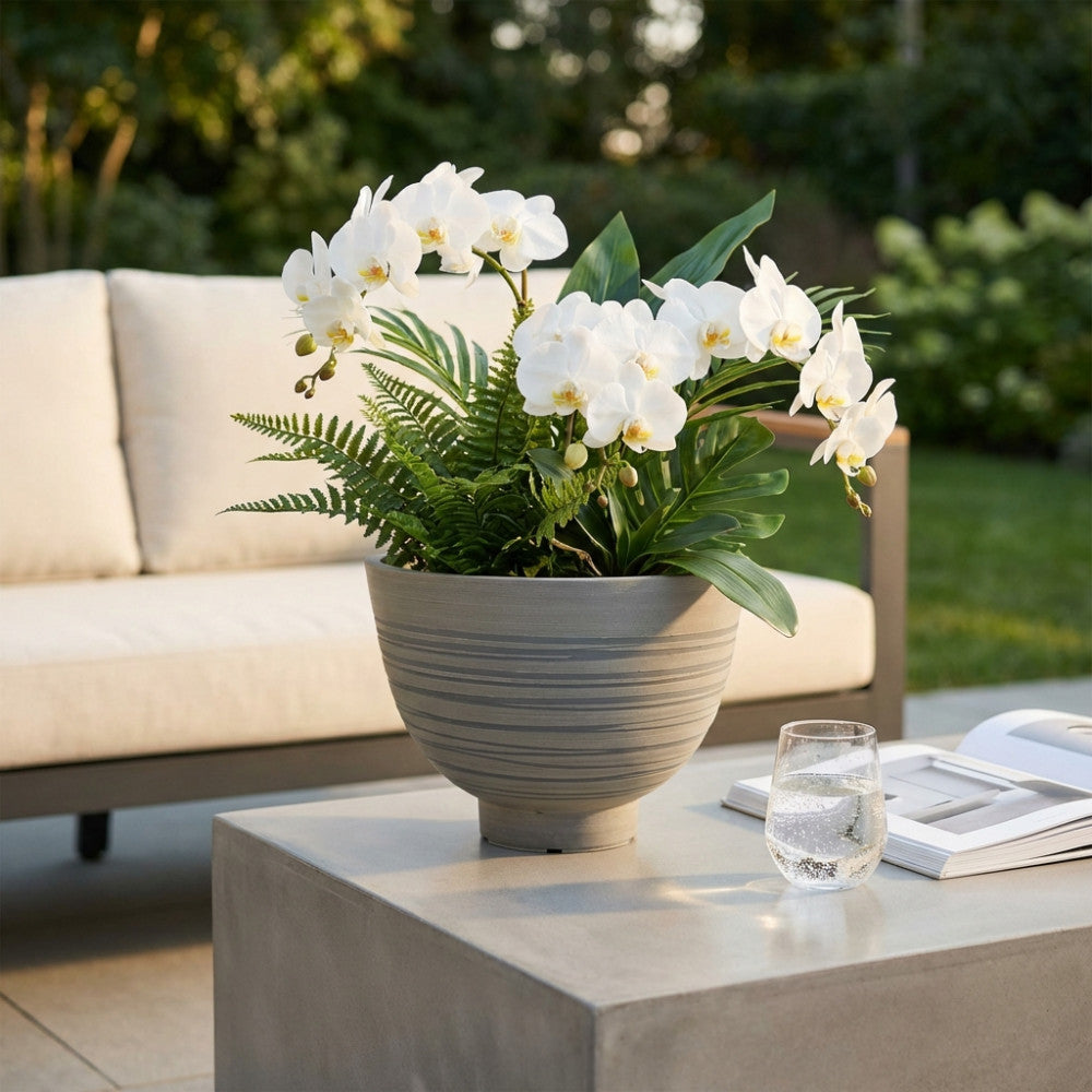 Vaso Grande Narciso in fibra sintetica antracite, decorato con orchidee bianche, posizionato su un tavolino in cemento all'aperto.