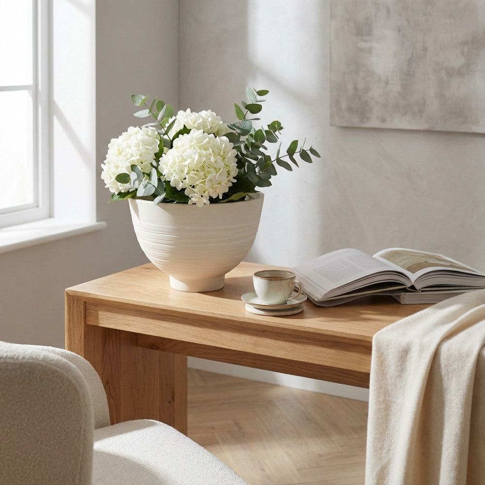 Vaso scultoreo bianco decorato con ortensie e rami di eucalipto su un tavolo in rovere chiaro.