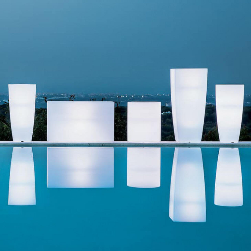 Composizione di vasi luminosi di diverse forme geometriche riflessi sull'acqua di una piscina al crepuscolo.