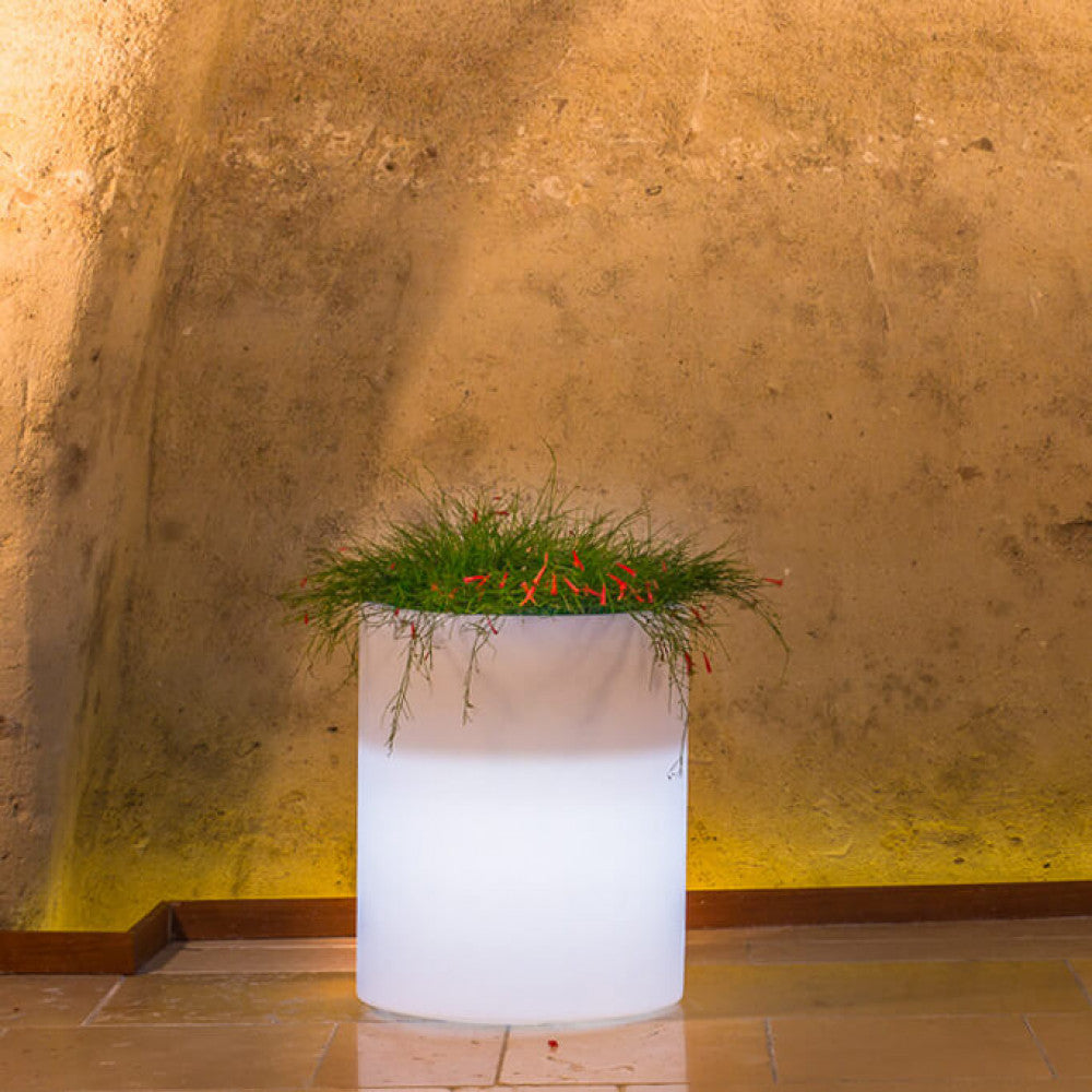 Vaso luminoso cilindrico acceso con luce bianca calda in un ambiente rustico con pareti in pietra.