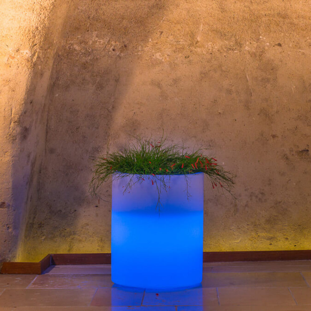 Vaso luminoso cilindrico acceso con luce blu intensa, inserito in un contesto architettonico in pietra.
