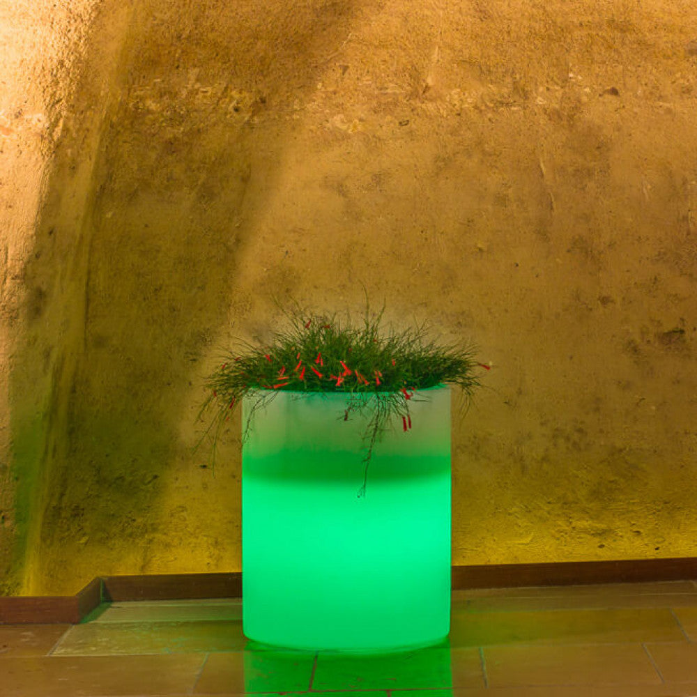 Vaso luminoso cilindrico acceso con luce verde smeraldo, posizionato su un pavimento in cotto.