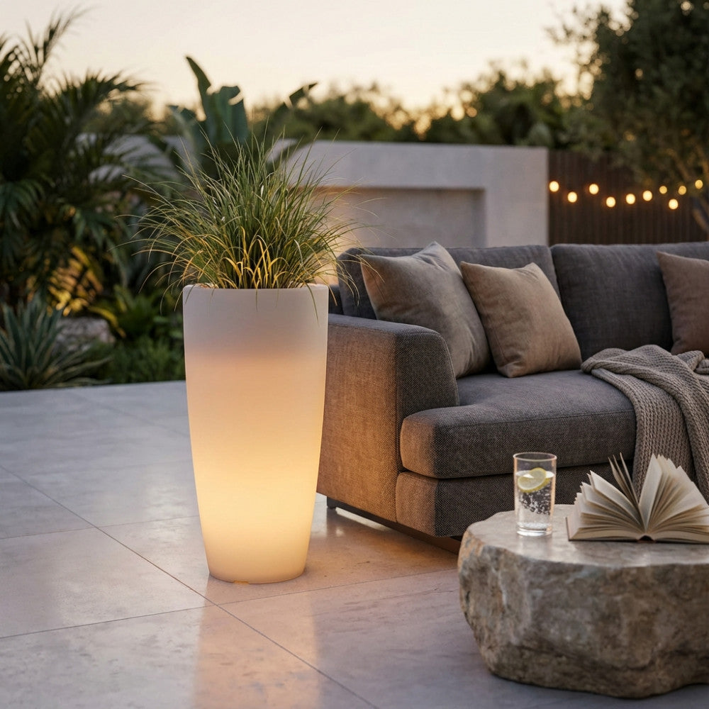 Vaso luminoso in polipropilene bianco acceso con luce calda, posizionato su un patio moderno accanto a un divano grigio.