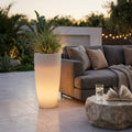 Vaso luminoso in polipropilene bianco acceso con luce calda, posizionato su un patio moderno accanto a un divano grigio.