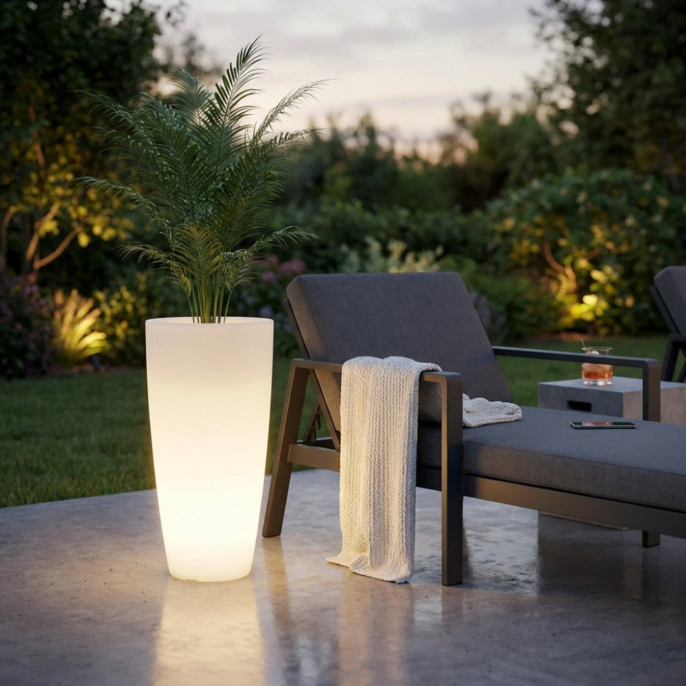 Fioriera illuminata Hydra con luce calda in un giardino serale, affiancata a un lettino prendisole di design.