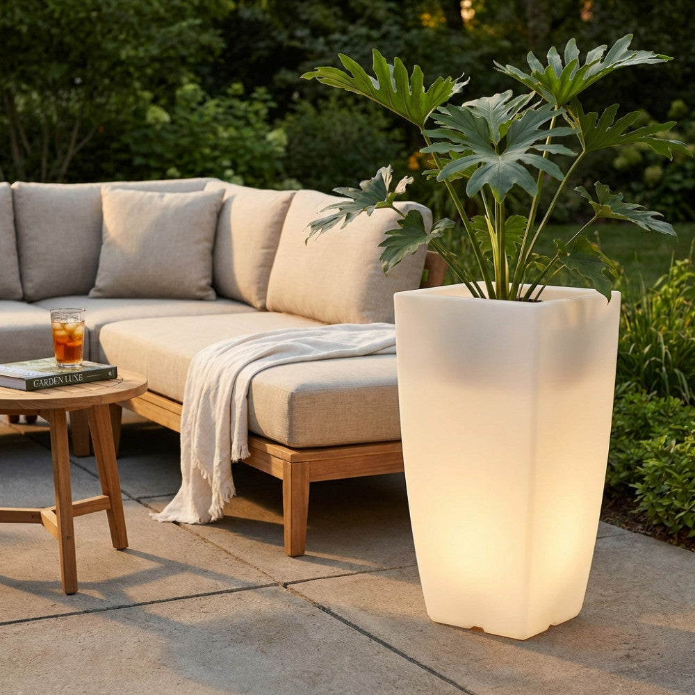 Vaso luminoso Hydrus acceso con luce bianca calda, posizionato su un patio in pietra accanto a un divano outdoor in tessuto chiaro.