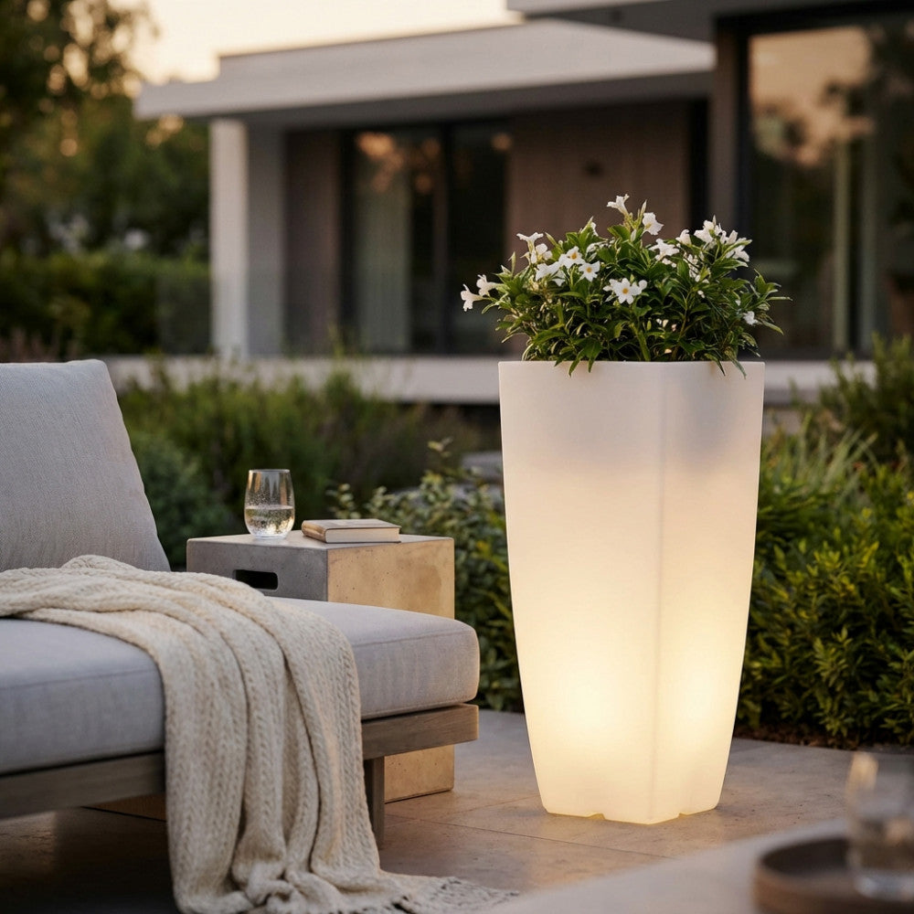 Vaso luminoso in polipropilene bianco acceso, decorato con fiori bianchi, situato in un giardino moderno al tramonto.