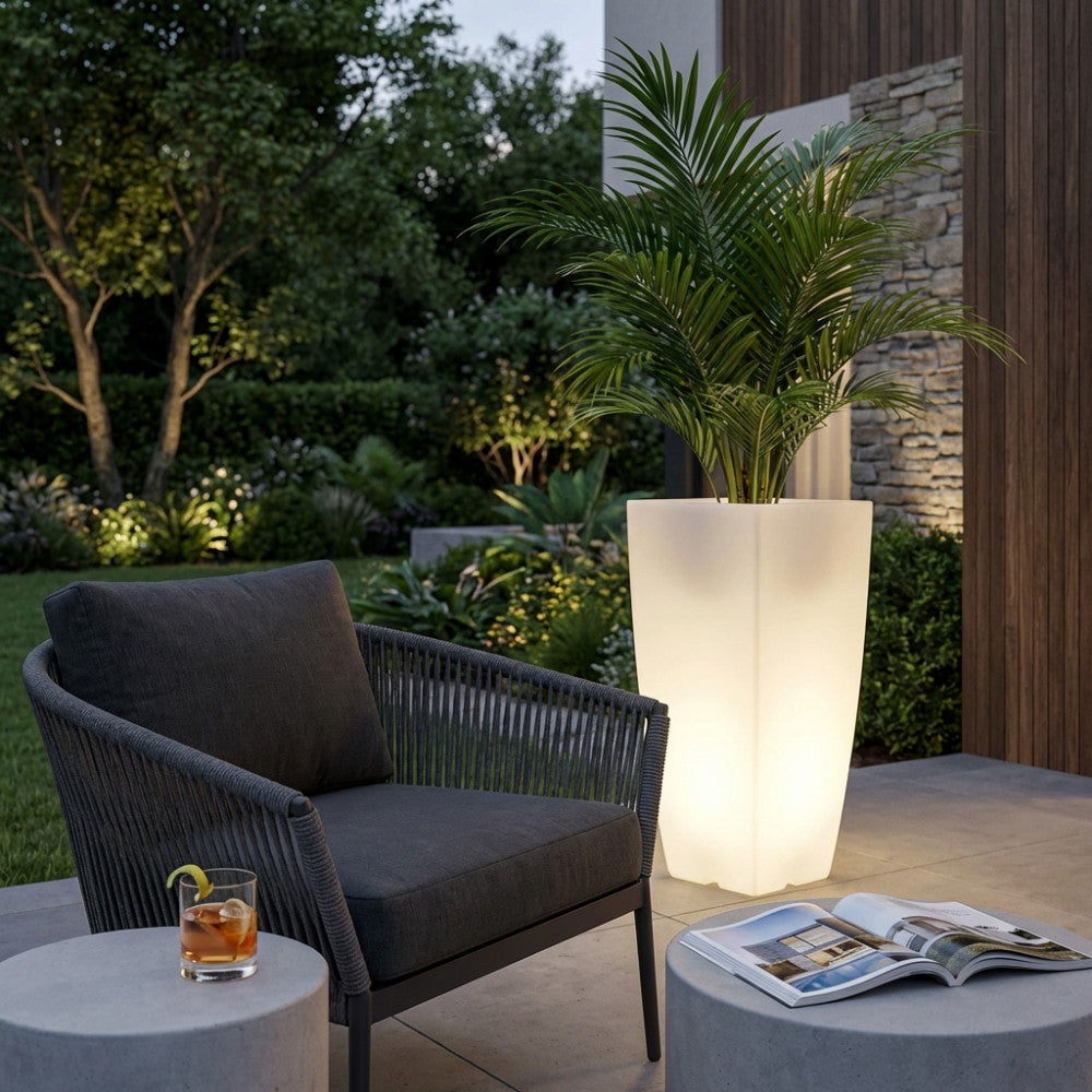 Vaso luminoso in polipropilene bianco posizionato all'aperto accanto a una poltrona in corda scura, illuminando una palma al tramonto.