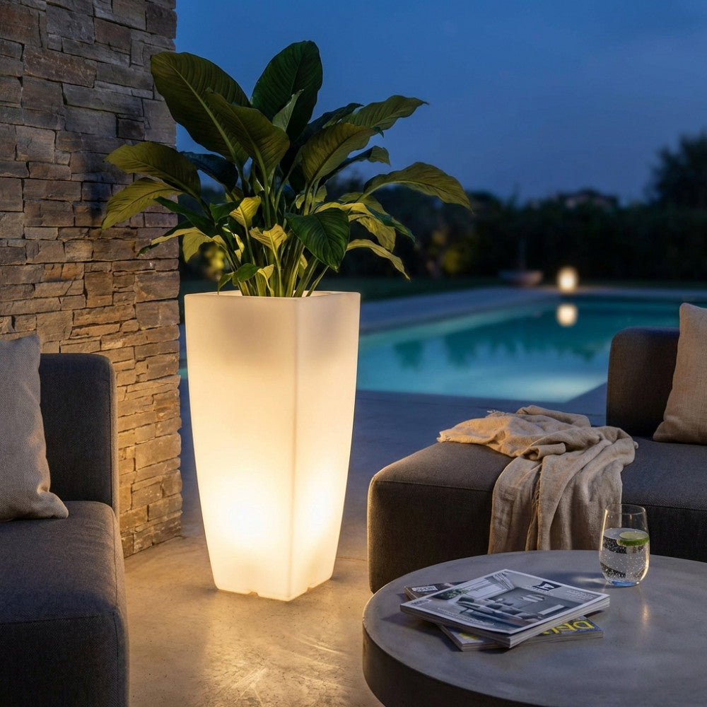 Vaso luminoso alto a bordo piscina durante la sera, che diffonde una luce calda accanto a un divano grigio elegante.