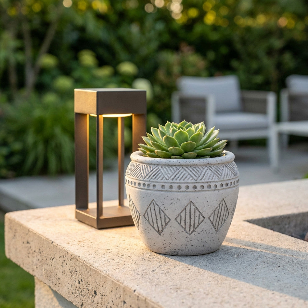 Vaso scultoreo grigio con pianta grassa, ambientato in un giardino esterno su un muretto in cemento accanto a una lanterna moderna.