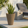 Vaso Medio Iris color taupe in fibra sintetica con pianta verde, posizionato su una terrazza in cemento accanto a un lettino prendisole.