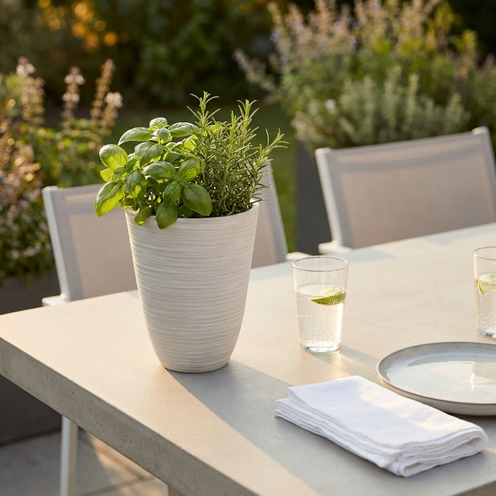 Vaso Piccolo Anemone bianco in fibra sintetica, posizionato su un tavolo da esterno in cemento, arricchito con piante aromatiche fresche.